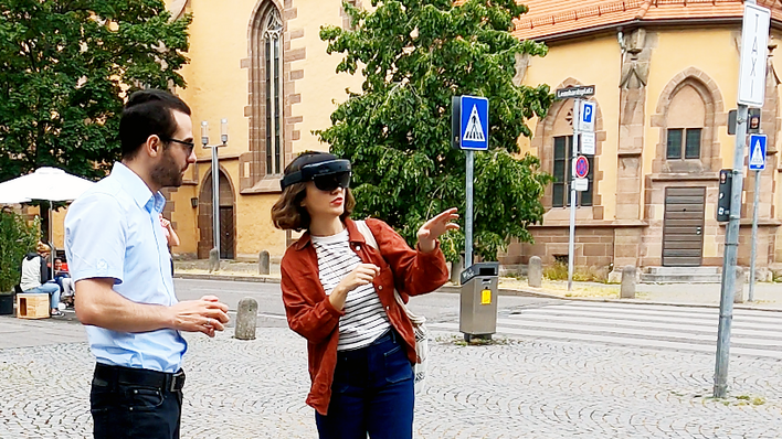 Eine Frau steht mit einer VR-Brille in der Leonhardsvorstadt und lässt sich von einem Mann die Anwendung erklären, im Hintergrund die Leonhardskirche